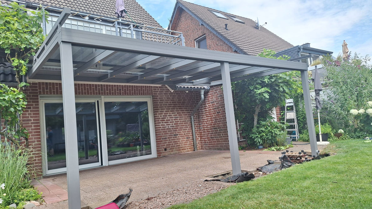 Terrassenüberdachung aus Aluminium in Grau mit klarem Glasdach und LED-Spots an einem Klinkerhaus mit Garten